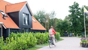 A person riding a bicycle on a paved path next to a building with a red-tiled roof and a dark facade. - Skylgerduin (Midsland)