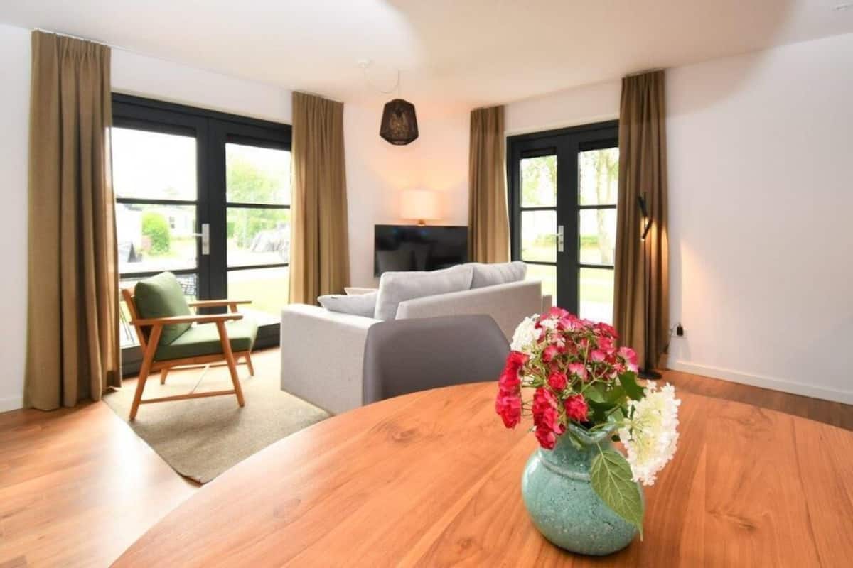 Una sala de estar moderna con una mesa redonda de madera, un sofá gris y una silla verde. Un televisor está montado en la pared, y hay un jarrón con flores sobre la mesa.