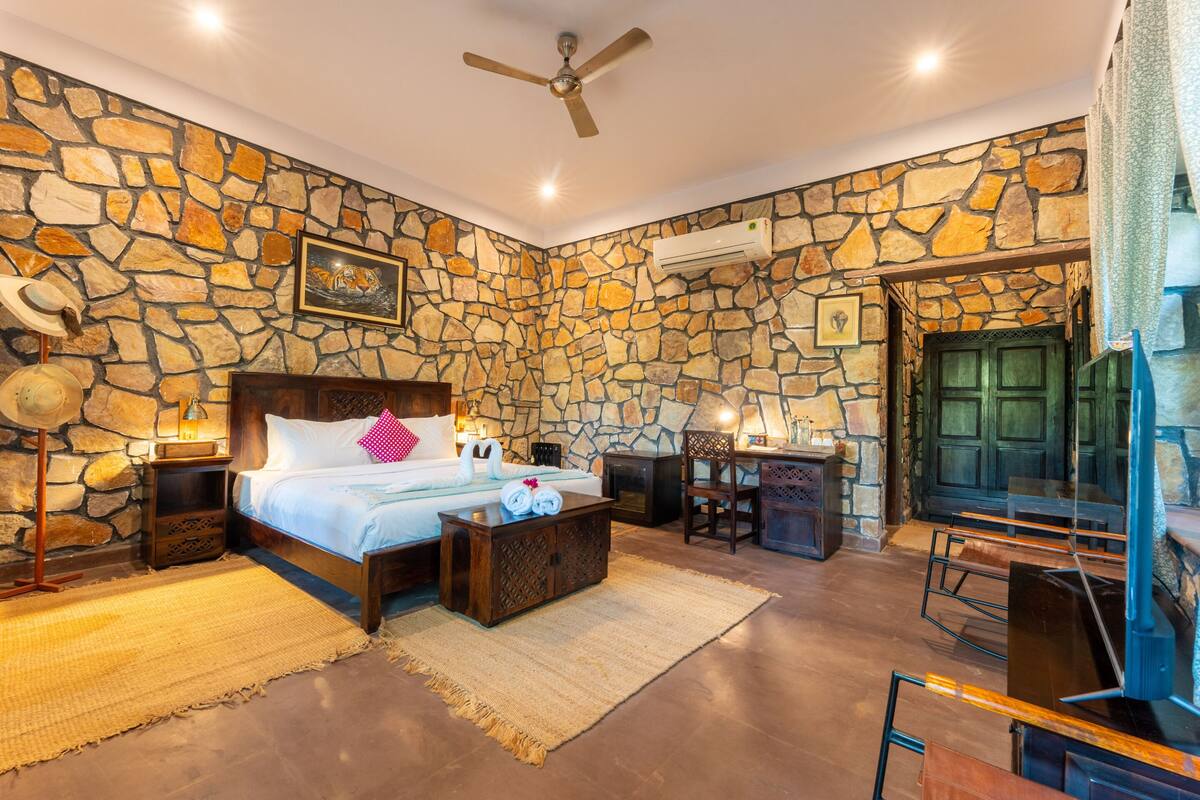A bedroom with a stone wall, a bed, a wooden nightstand, a desk, and a chair.