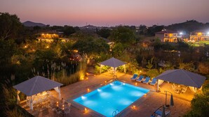 Piscina com espreguiçadeiras e guarda-sóis, cercada por árvores e vista para o pôr do sol.