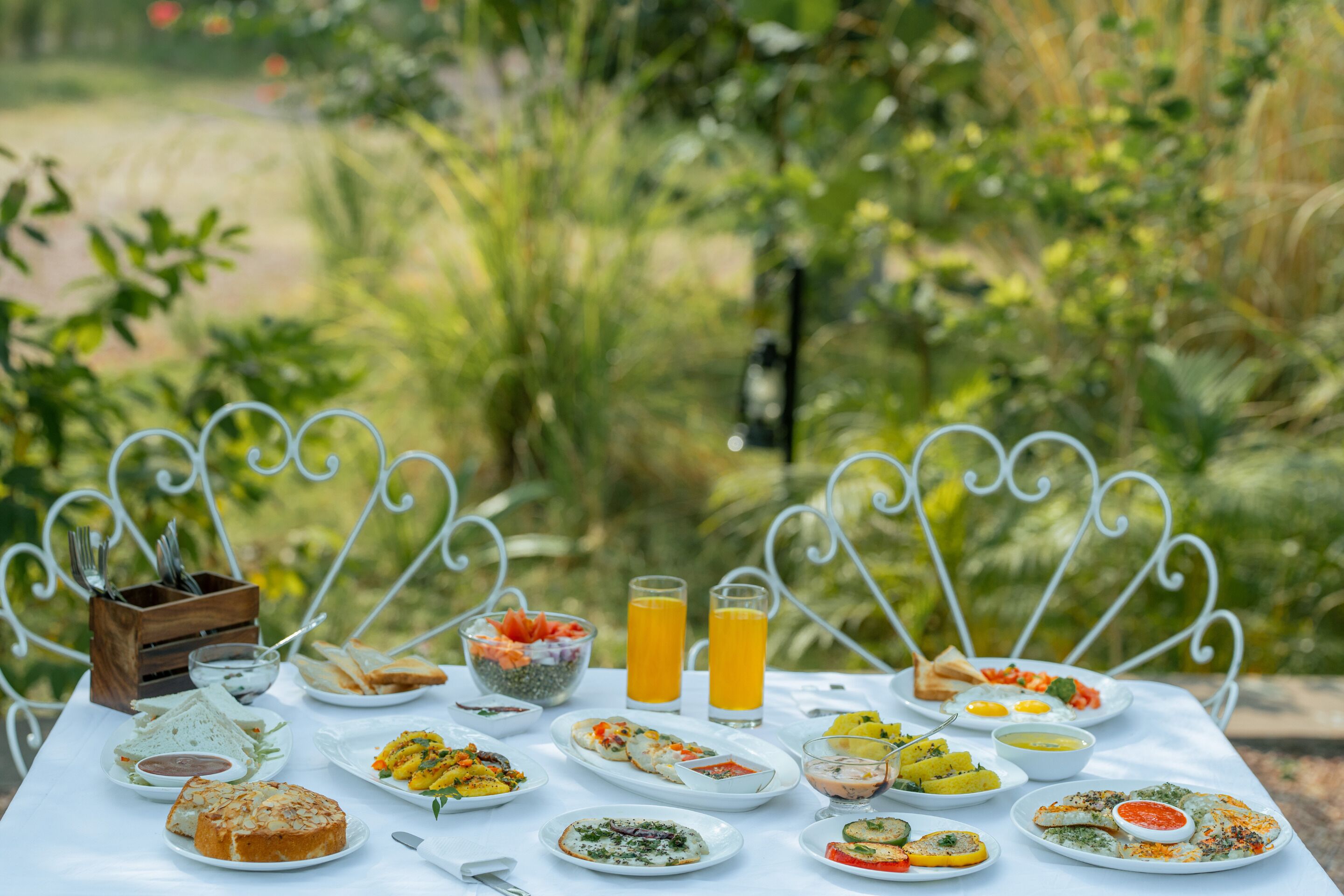 A table set outdoors with various dishes and drinks, surrounded by greenery.