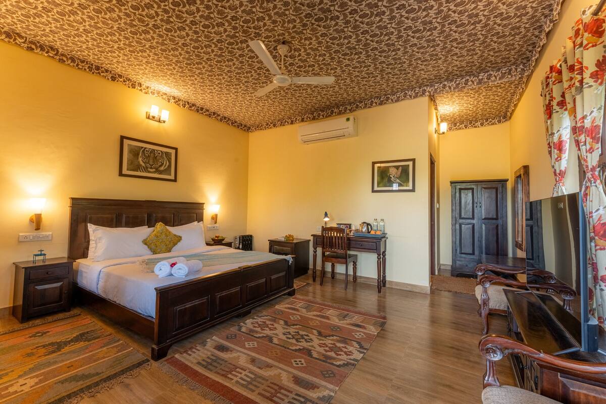 A bedroom with a patterned ceiling, a bed with pillows, a desk with a chair, and a wooden floor.