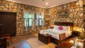 Quarto com parede de pedra, cama com roupa branca, cabeceira de madeira, mesa de cabeceira com abajur e janela com cortinas.