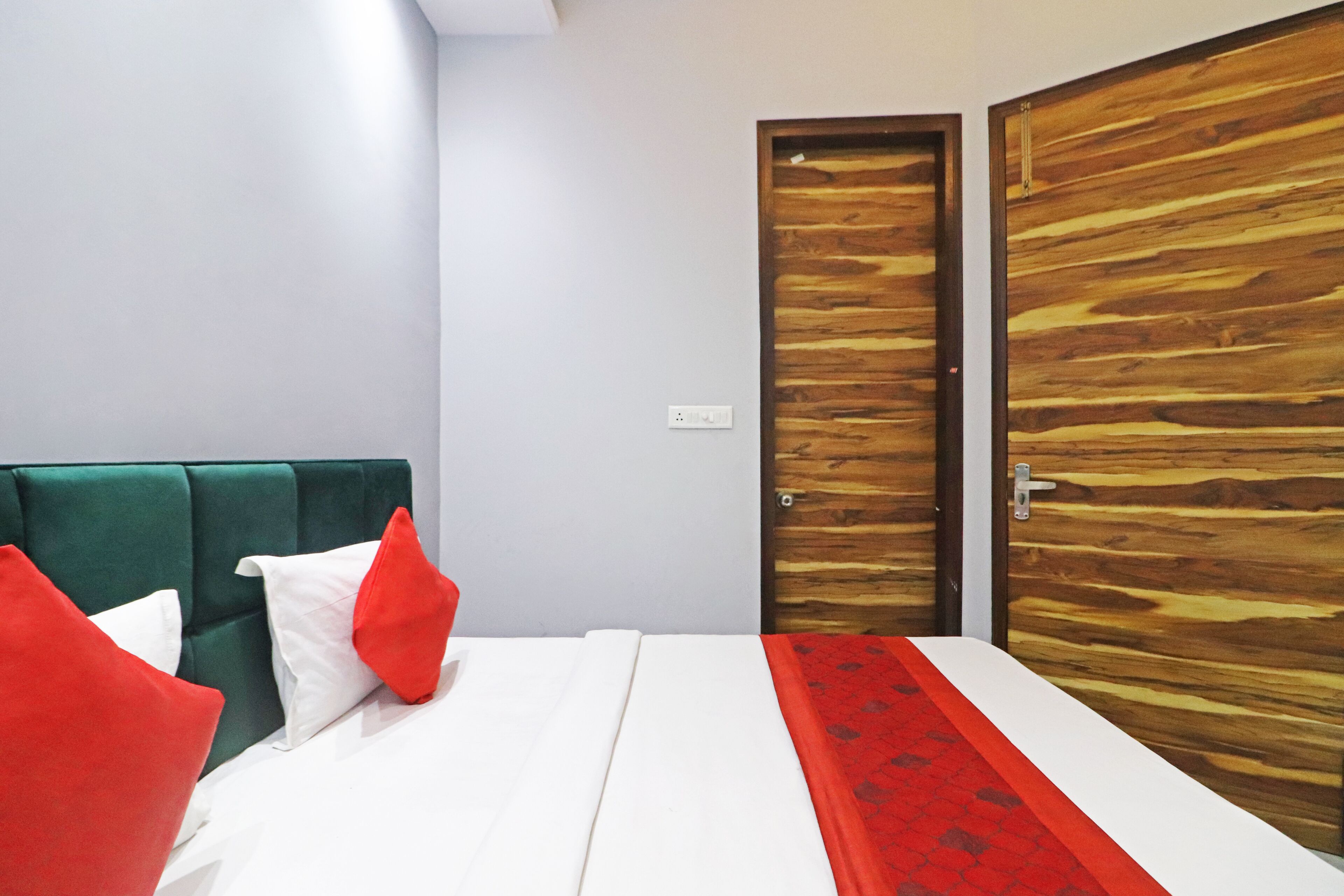 Habitación de hotel con cama, cabecera tapizada en verde, almohadas rojas y blancas, una puerta de madera y un corredor de cama con estampado rojo.