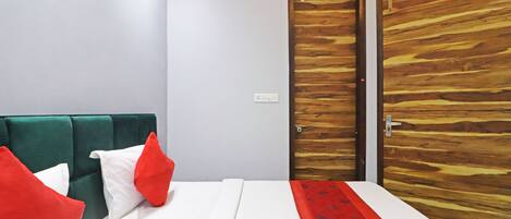 Habitación de hotel con cama, cabecera tapizada en verde, almohadas rojas y blancas, una puerta de madera y un corredor de cama con estampado rojo.
