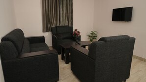 A living room with dark grey sofas, a small table with a vase of flowers, a flat-screen TV mounted on the wall, and a window with curtains. - Srushti Service Apartment (Bengaluru)