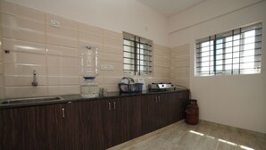 A kitchen with dark wooden cabinets, a large water dispenser, a gas cylinder, and a window with bars. - Srushti Service Apartment (Bengaluru)