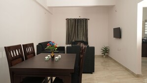 A dining area with a wooden table and chairs, a vase with flowers, and a grey sofa. - Srushti Service Apartment (Bengaluru)