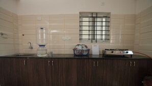 A kitchen with a water dispenser, a window with a metal grid, and a gas stove. - Srushti Service Apartment (Bengaluru)