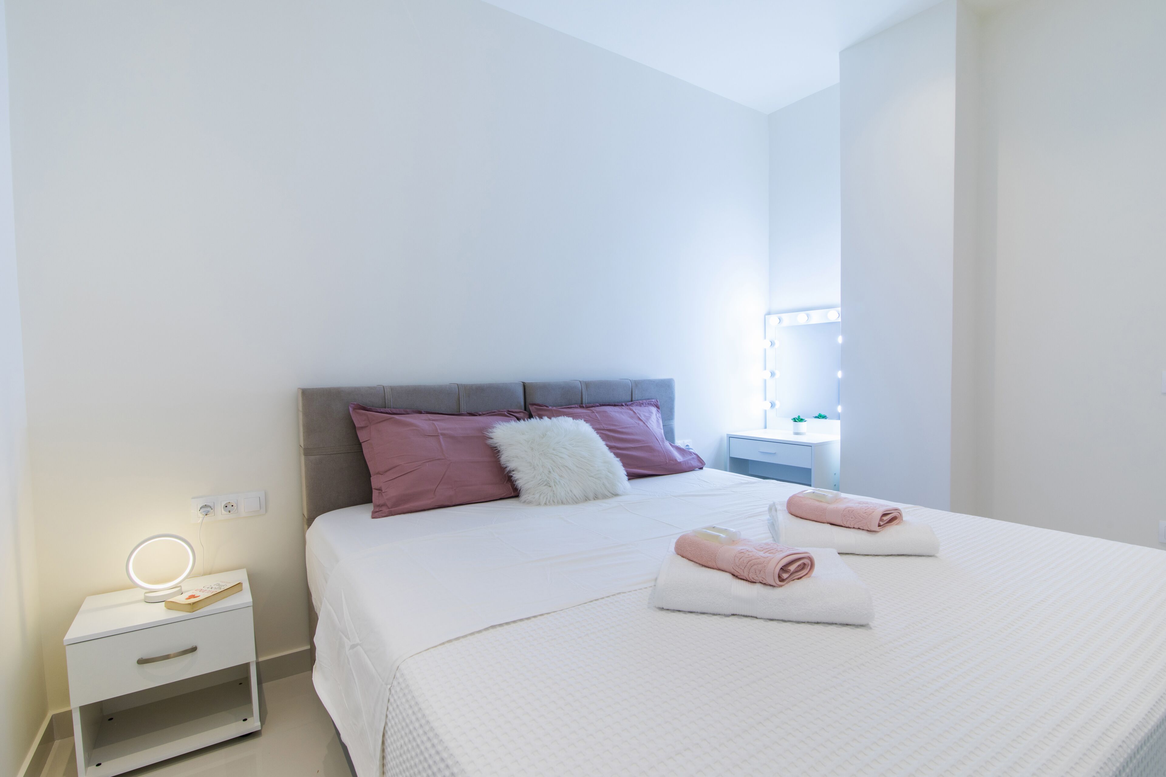 A neatly made bed with pink and white pillows, a bedside table with a mirror, and two towels.