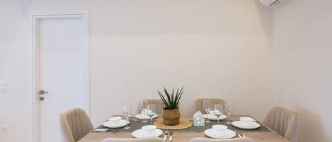 Een eethoek met een tafel gedekt voor vier personen, inclusief borden, glazen en bestek. In het midden staat een potplant, en de kamer heeft een witte deur en een airconditioning op de muur.