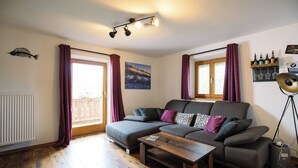 A living room with a grey sofa, a wooden coffee table, and a floor lamp. There is a window with curtains, a radiator, and a wall-mounted shelf with bottles.