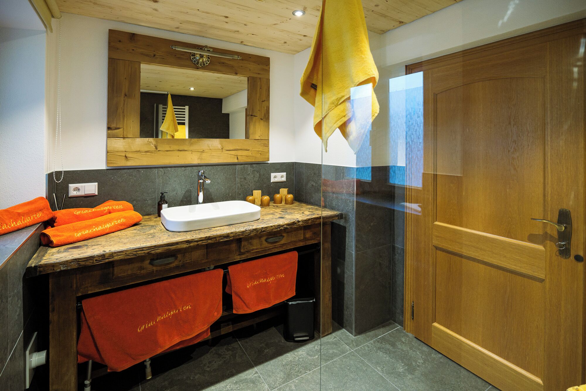Un baño con tocador de madera, un lavabo blanco y un espejo encima. Hay toallas colgadas en la pared y una puerta de madera.