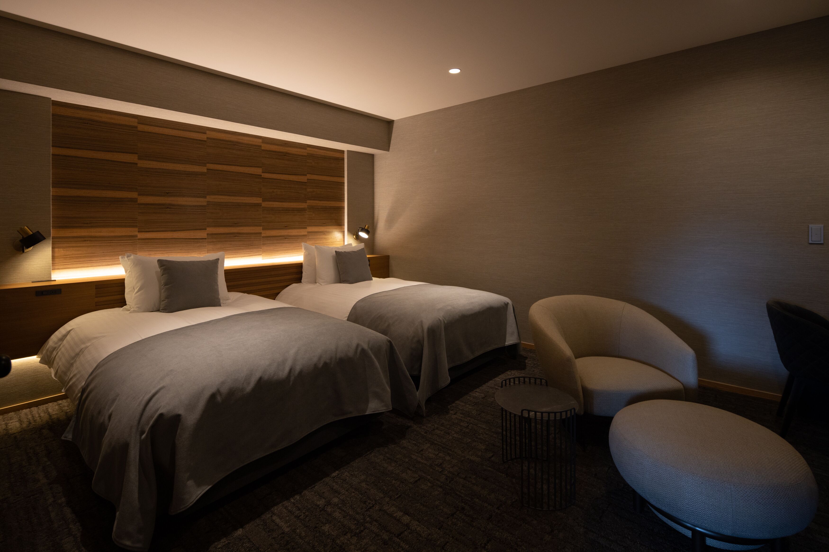 A modern hotel room with two beds, a curved armchair, a small round table, and a wooden headboard.