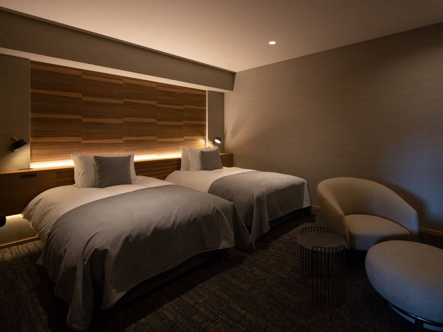 A modern hotel room with two beds, a curved armchair, a small round table, and a wooden headboard.