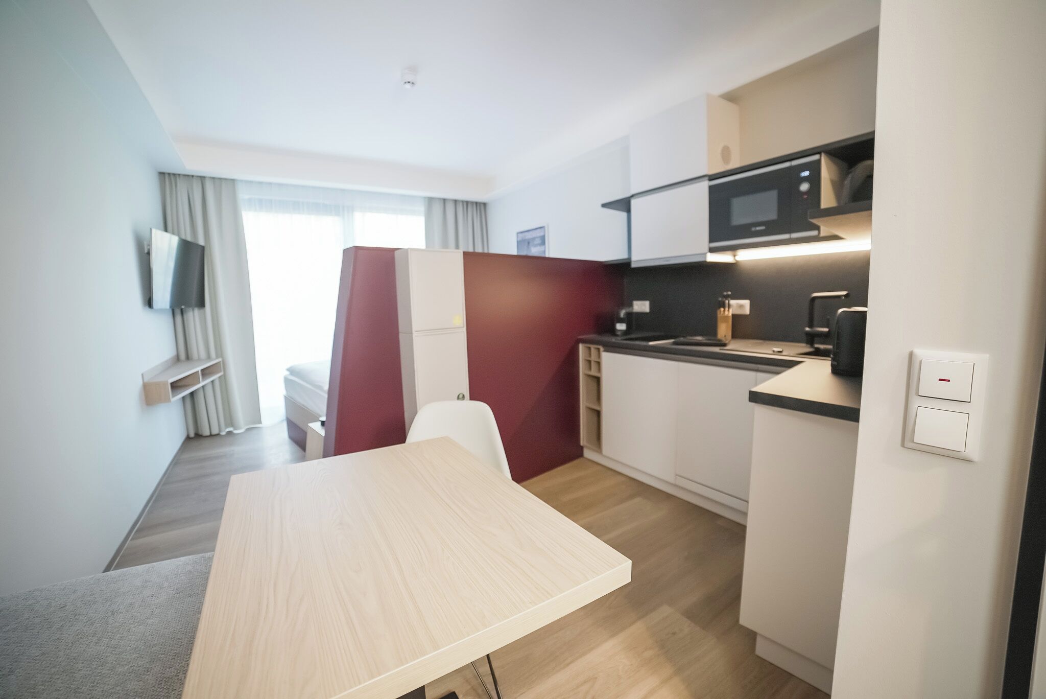 A compact hotel room with a kitchenette, a dining area, and a TV mounted on the wall.