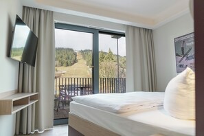 Une chambre d’hôtel avec un lit, un téléviseur fixé au mur, un balcon donnant sur une forêt et une route, et une affiche accrochée au mur.