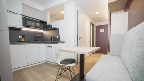 Une cuisine de taille réduite avec des meubles blancs, un plan de travail noir, une chaise blanche et un canapé gris.
