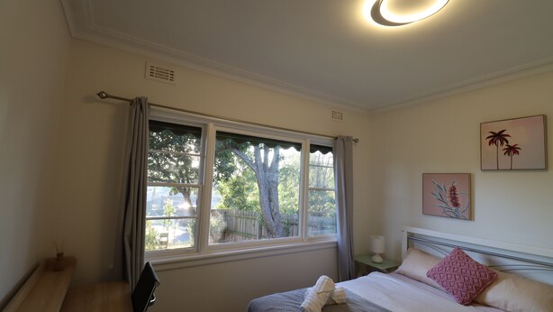 A bedroom with a bed, a desk, a window with curtains, a nightstand, and a wall-mounted lamp. - Humble Abode near Train Station, Costco, Eastland (Heathmont)