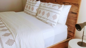 Een net opgemaakt bed met witte lakens en gestreepte kussens, een houten hoofdbord, een rond nachtkastje met een lamp, en een spiegel aan de muur.