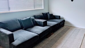 Een moderne woonkamer met een hoekbank, een groot raam met zonwering en een geweven vloerkleed.