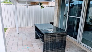 Een terras met een tafel met glazen blad, rieten meubels en een schuifpui die leidt naar een overdekt gedeelte.