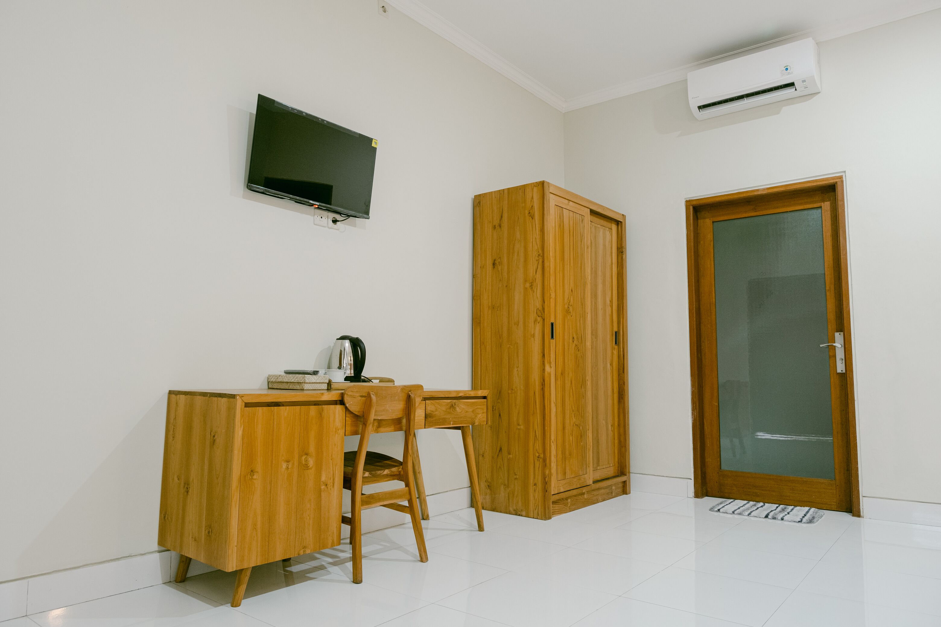 A room with a wooden desk, chair, and wardrobe, a television mounted on the wall, and a door with a glass panel.