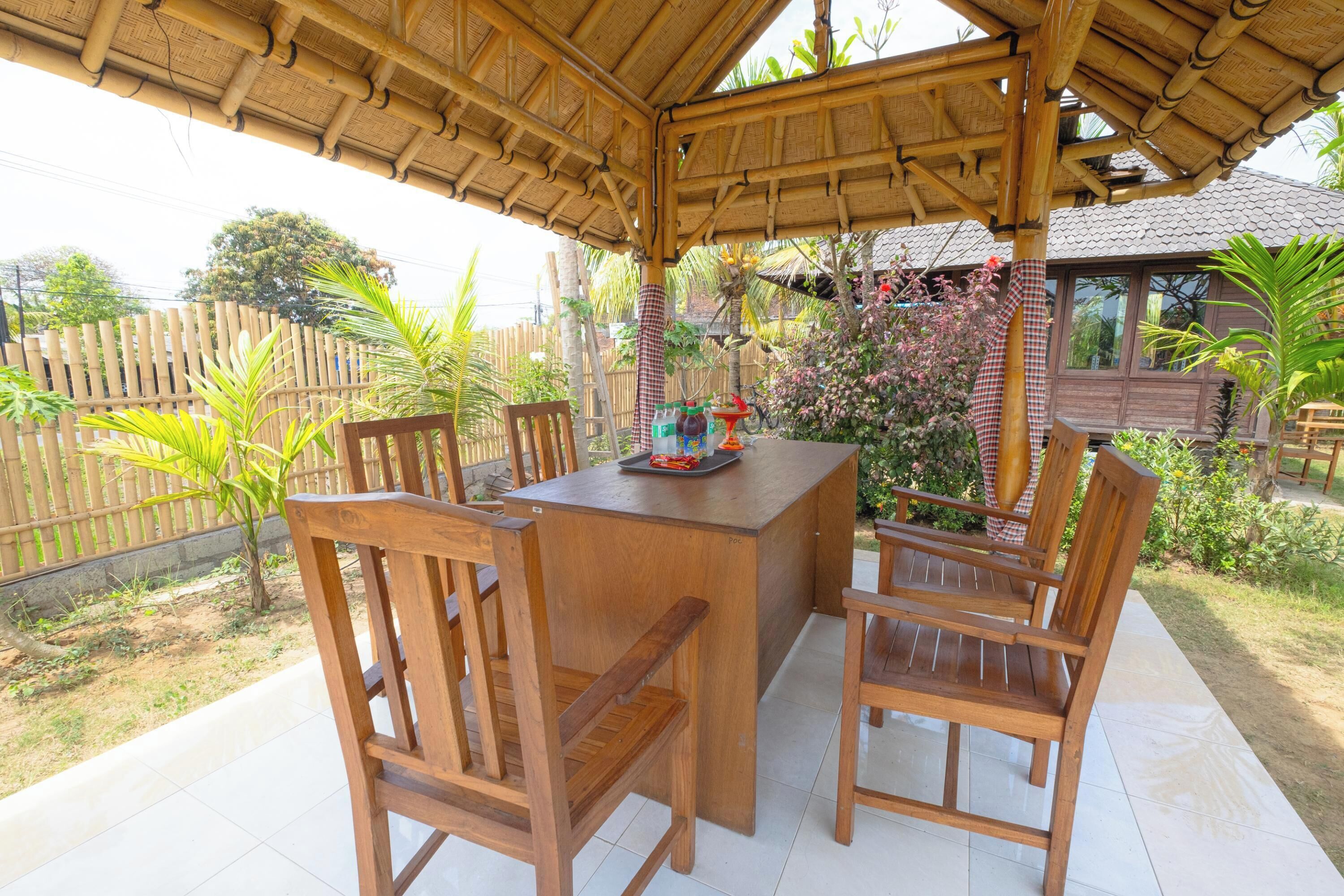 An outdoor wooden table and chairs under a bamboo roof, surrounded by plants and a wooden fence.