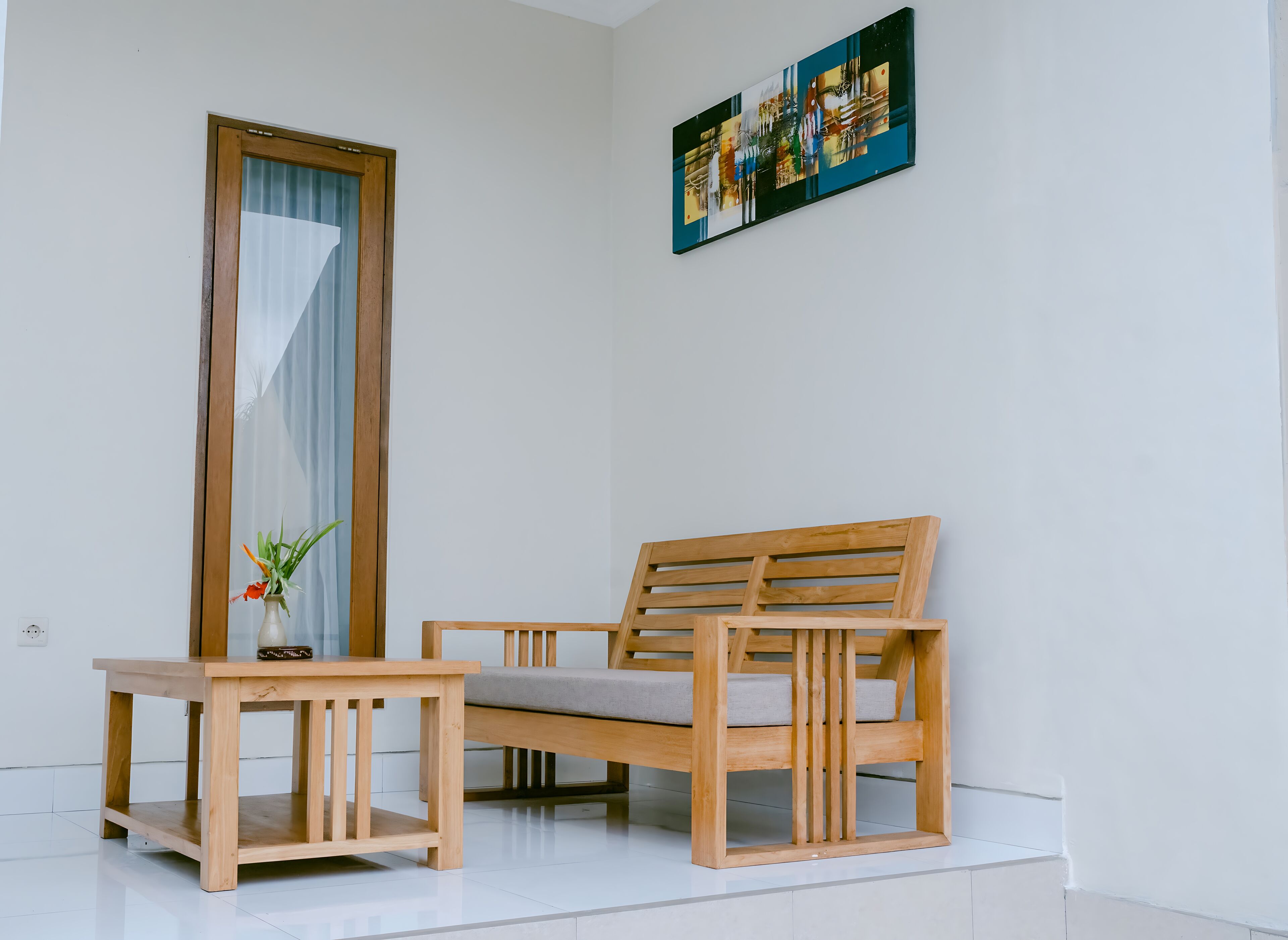A wooden bench with a cushion, a small wooden table with a vase of flowers, and a painting on the wall.