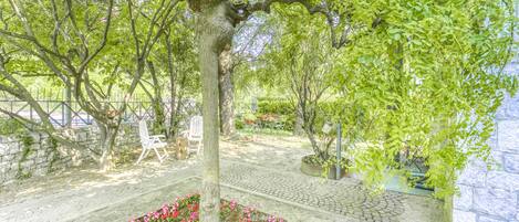 A garden with a tree, a flower bed, and a paved area with chairs and tables.