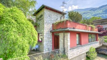 Un bâtiment en pierre avec une partie supérieure rouge, une petite tour surmontée d’une antenne et un jardin agrémenté de plantes en pot.