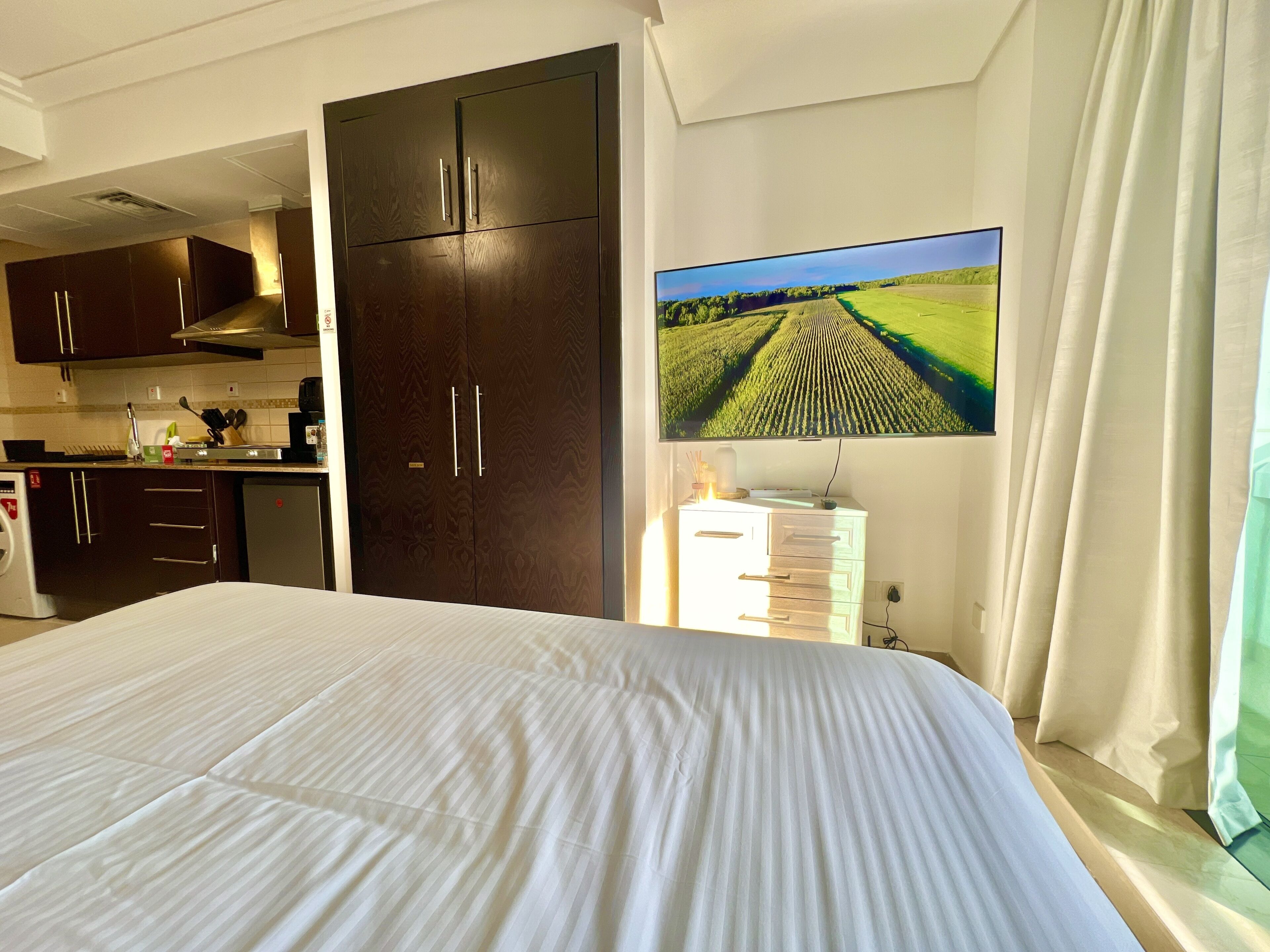 A bedroom with a large bed, a TV mounted on the wall, and a kitchen area in the background.