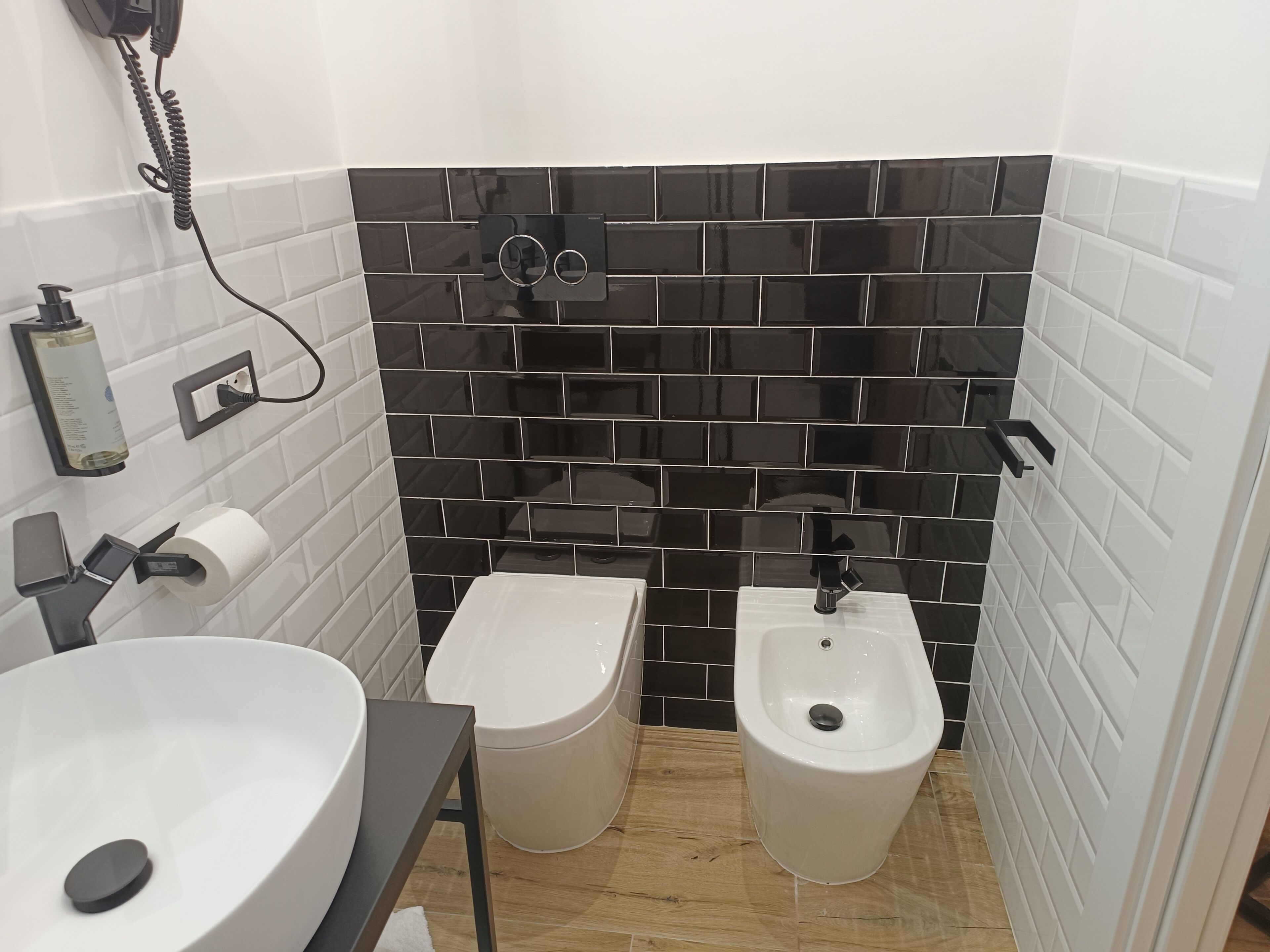 Casa de banho compacta com lavatório branco, duas sanitas, um bidé e um painel de azulejos pretos.