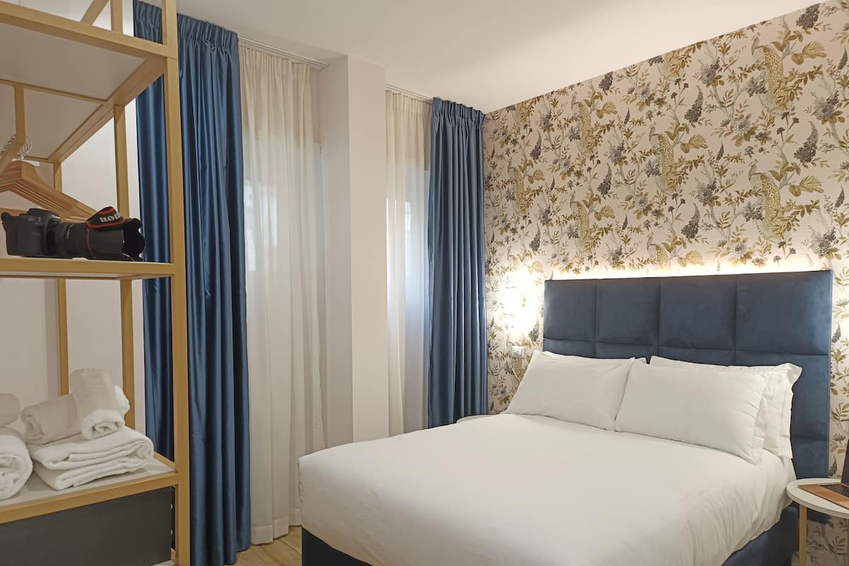 A neatly made bed with a blue headboard, a wooden shelf with folded towels, and a wall with floral wallpaper.