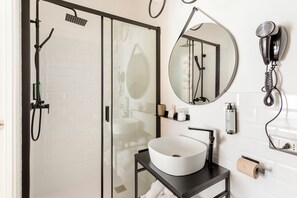 A modern bathroom with a glass shower enclosure, a round mirror, a white sink, and a hairdryer mounted on the wall. - Romi Suite (Rome)