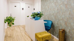 Un espace d’accueil avec un bureau blanc, une chaise bleue, un pouf jaune et une plante en pot.