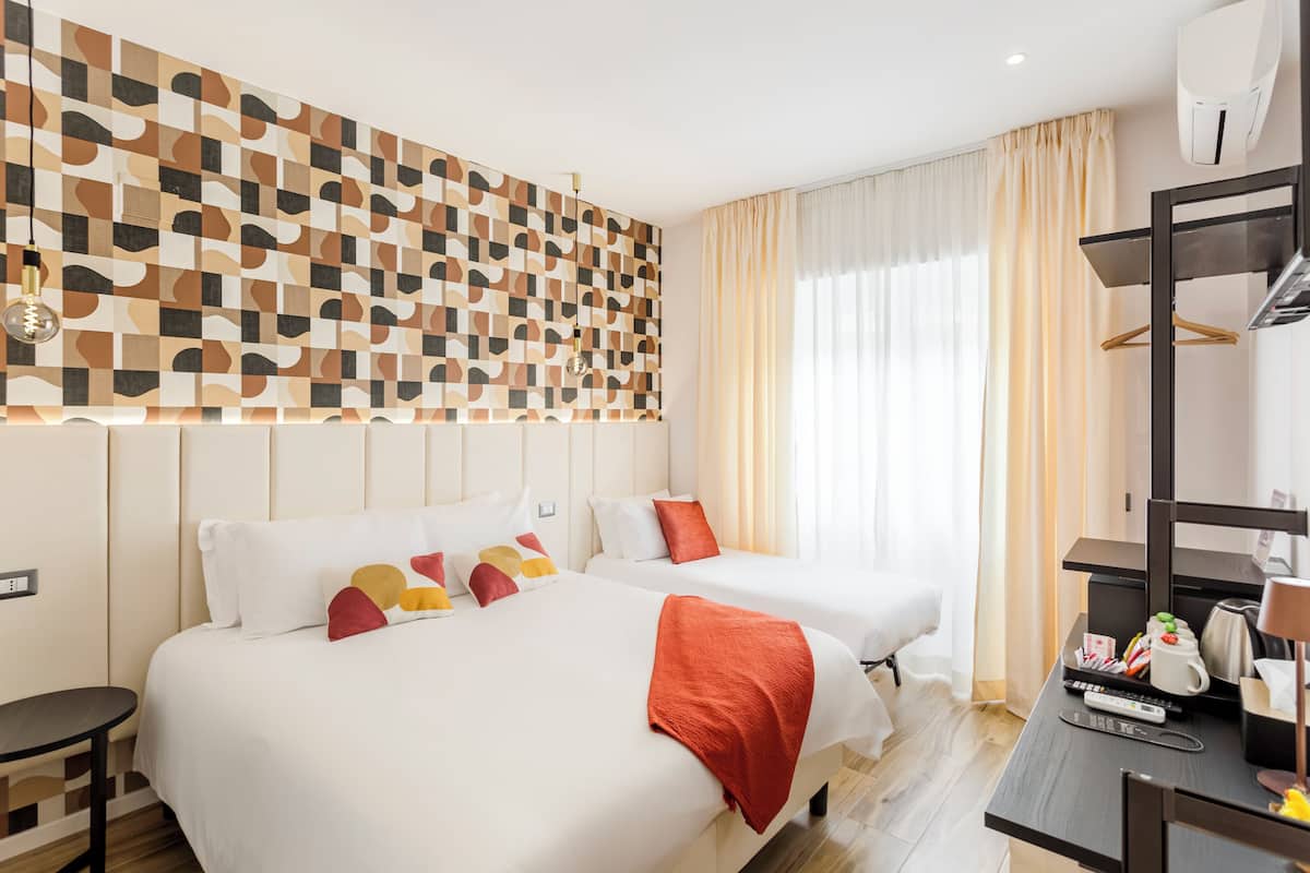 A hotel room with two beds, a desk with a computer and a coffee maker, a wall-mounted air conditioner, and a patterned wall.