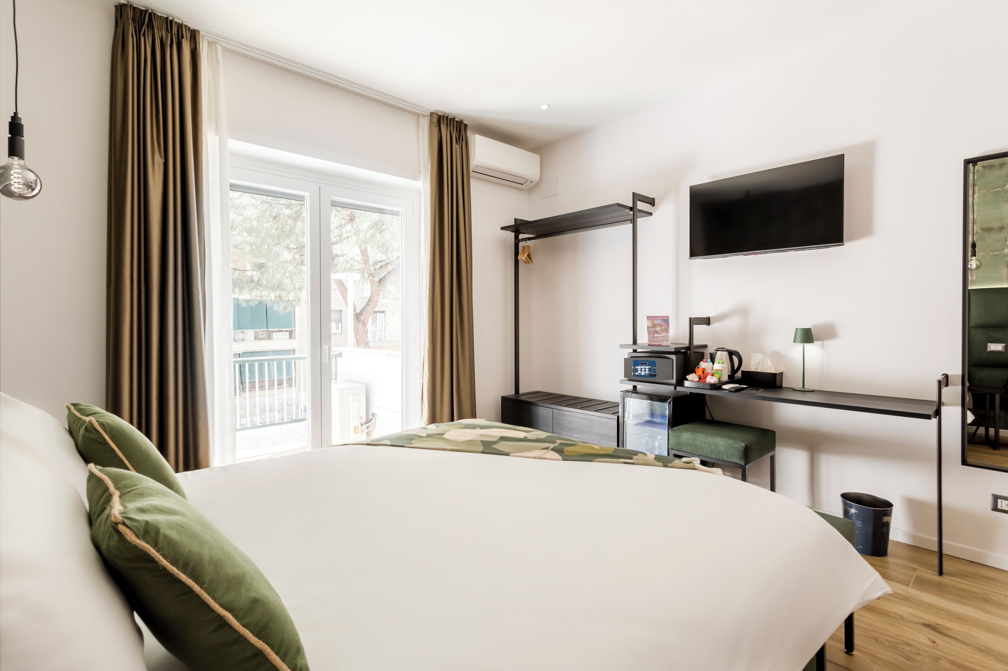Habitación de hotel con una cama grande, un escritorio con televisor de pantalla plana, una pequeña cocina americana y un balcón con cortinas.