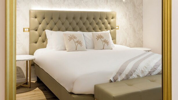 Une chambre moderne avec une tête de lit capitonnée, un grand lit avec une literie blanche, un banc au pied du lit et deux tables de chevet.