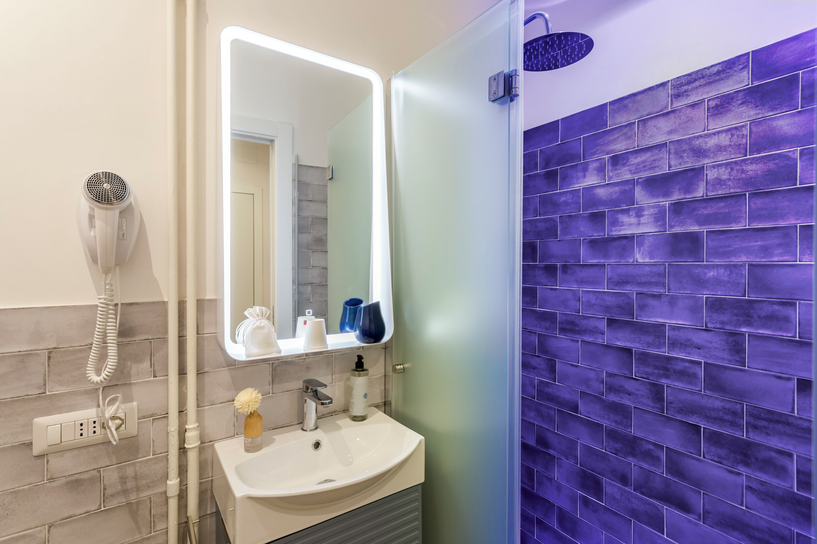A bathroom with a white sink, a mirror, a hairdryer, and a shower with purple tiles.