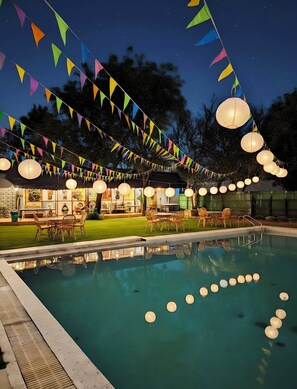 A pool area with a clear view of the pool, surrounded by tables and chairs, and decorated with colorful triangular flags and hanging lanterns. - Chopra Farms and Resorts (Gurugram)