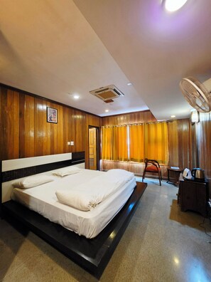 A hotel room with a large bed, a desk with a chair, a fan, and a small table with a kettle. - Chopra Farms and Resorts (Gurugram)