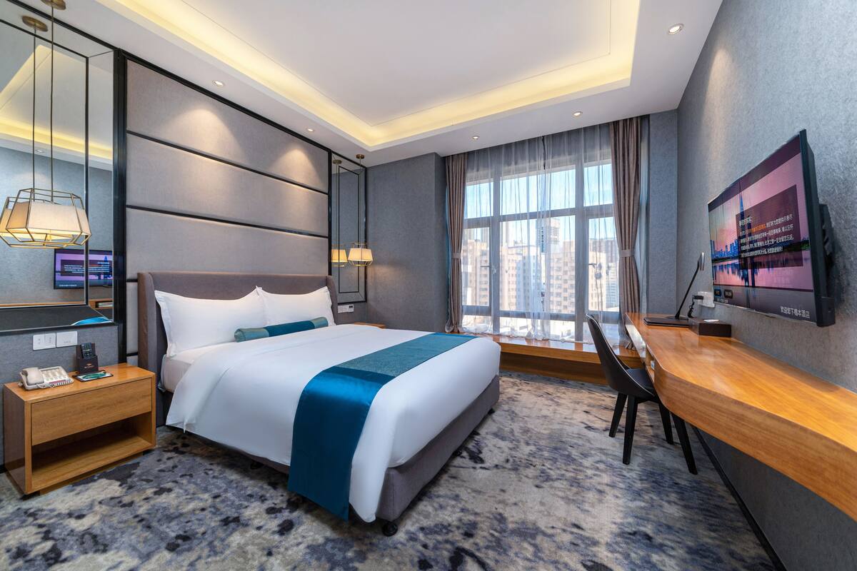 A modern hotel room with a large bed, bedside tables, a desk, and a TV on the wall.