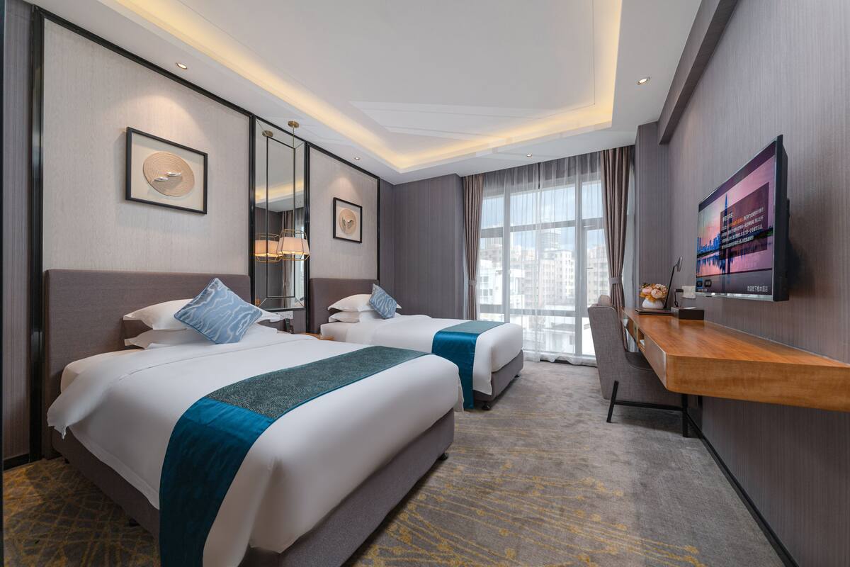 A hotel room with two beds, a desk, a TV, and a large window.