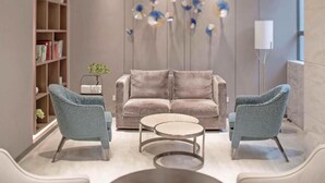 A modern living room with a beige sofa, two armchairs, and a coffee table. There is a bookshelf with books and decorative items, and a wall-mounted light fixture with blue and white flowers. - deluxe king (Shenzhen)