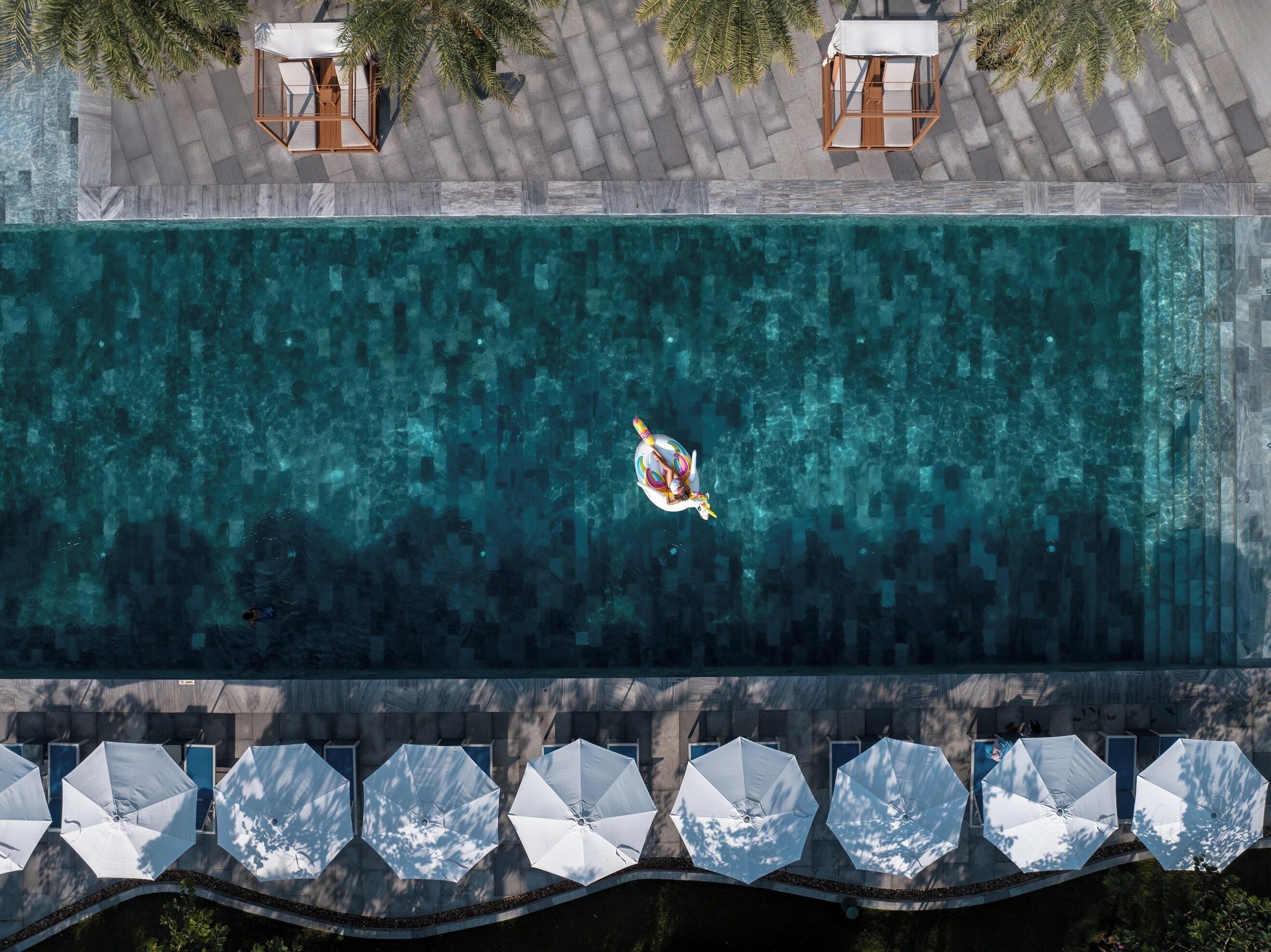 Vue aérienne d’une piscine où l’on aperçoit une personne flottant sur un flotteur, entourée de chaises longues et de parasols.