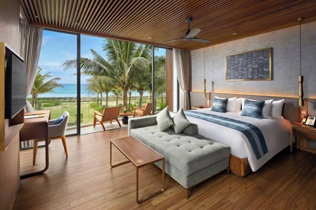 A modern hotel room with a large bed, a sofa, a desk, and a view of the ocean.