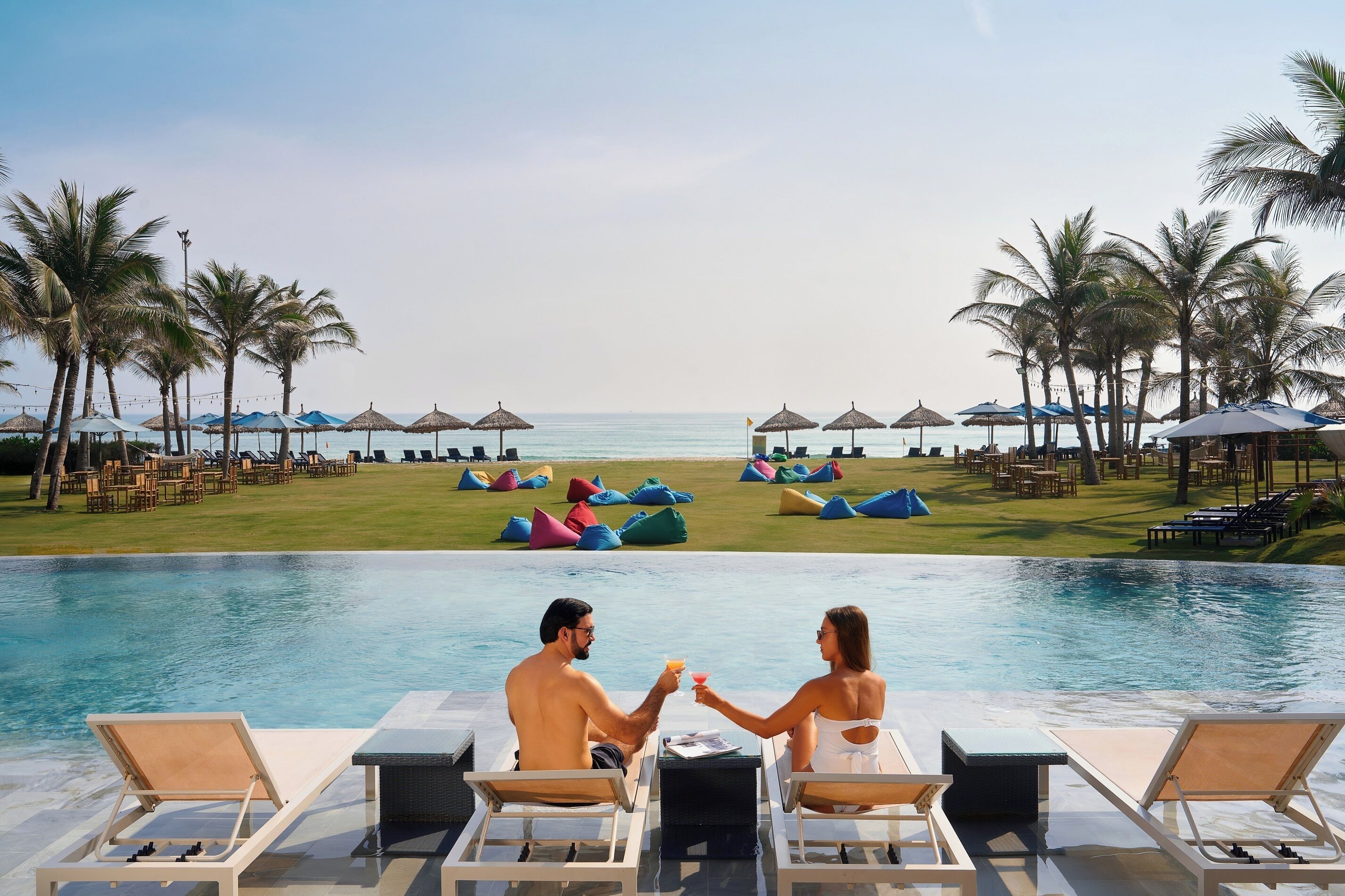 Una pareja junto a una piscina con sillones de descanso y sombrillas, palmeras y vista a la playa.