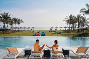 Una pareja junto a una piscina con sillones de descanso y sombrillas, palmeras y vista a la playa.