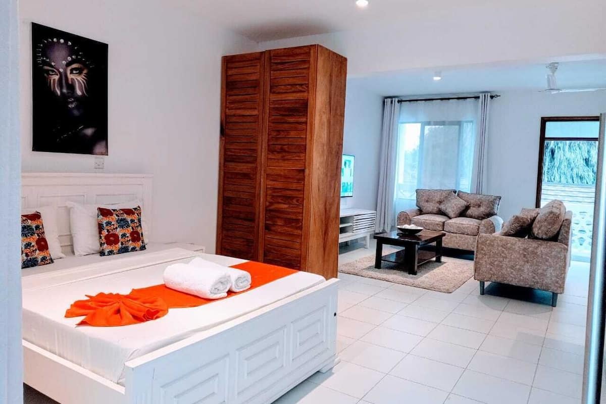 A modern hotel room with a large bed, a wooden wardrobe, a seating area with a sofa and armchairs, and a large window providing a view.